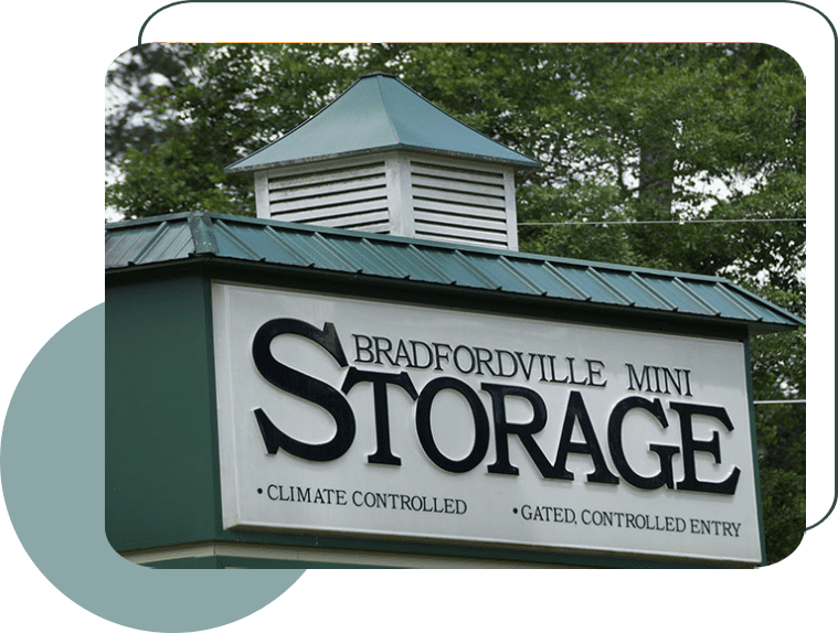 Sign for "Bradfordville Mini Storage" with a green roof, white background and black lettering. The sign also mentions "Climate Controlled" and "Gated, Controlled Entry." Trees are visible in the background.